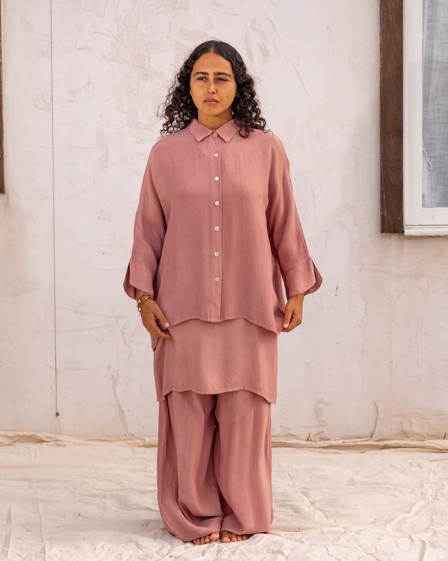 
                  
                    woman dressed in bouviet oversized shirt and miguel straight legged pants in orange peel fibre in dirty rose pink standing in front of a white wall
                  
                