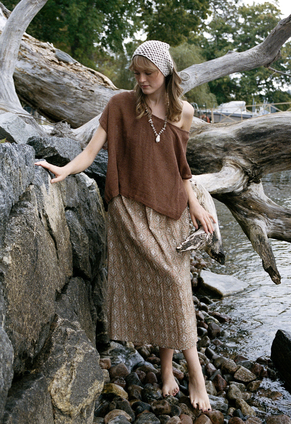 woman dressed in beige patterned midi dress with oversized net rust top in front of drift wood