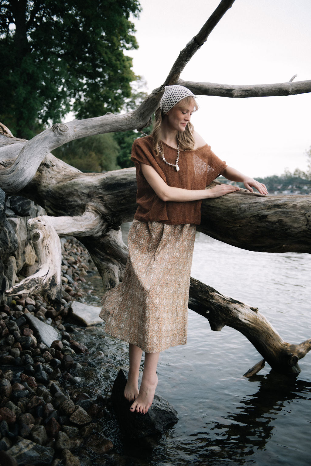 
                  
                    woman dressed in beige patterned midi dress with oversized net rust top in front of drift wood
                  
                