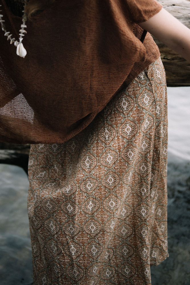 
                  
                    woman dressed in beige patterned midi dress with oversized net rust top in front of drift wood
                  
                