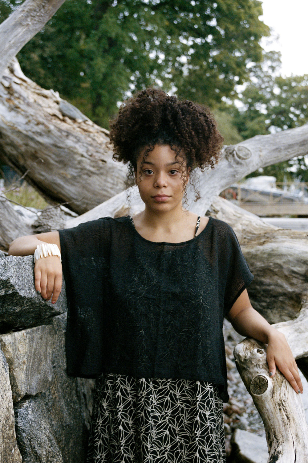 woman dressed in dark patterned mini dress with oversized net charcoal top in front of drift wood