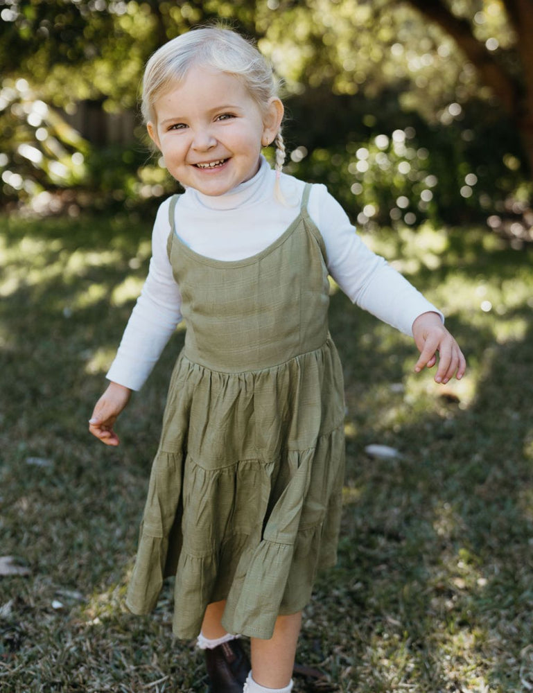 
                  
                    young girl dressed in vincente organic sugarcane tie dress in green forest standing on a lawn smiling
                  
                