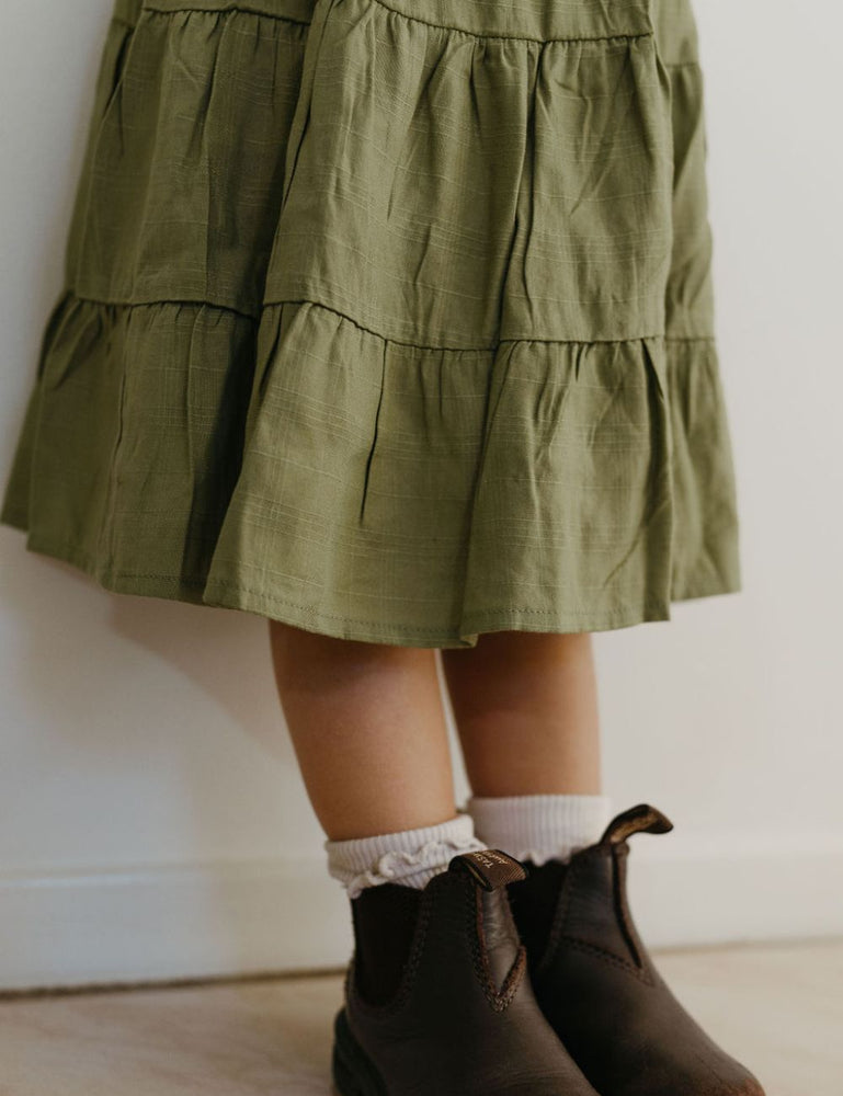 
                  
                    young girl dressed in vincente organic sugarcane tie dress in green forest standing in front of a white wall
                  
                